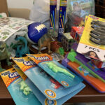 A table full of fidgets waiting to be packed and sent to the Scholars: glow bracelets, Nerf football, large plastic dinosaur, grow animals that you put in water, mustaches
