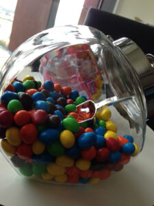 Clear glass candy jar with a metal lid, filled with peanut M&Ms and a candy scoop